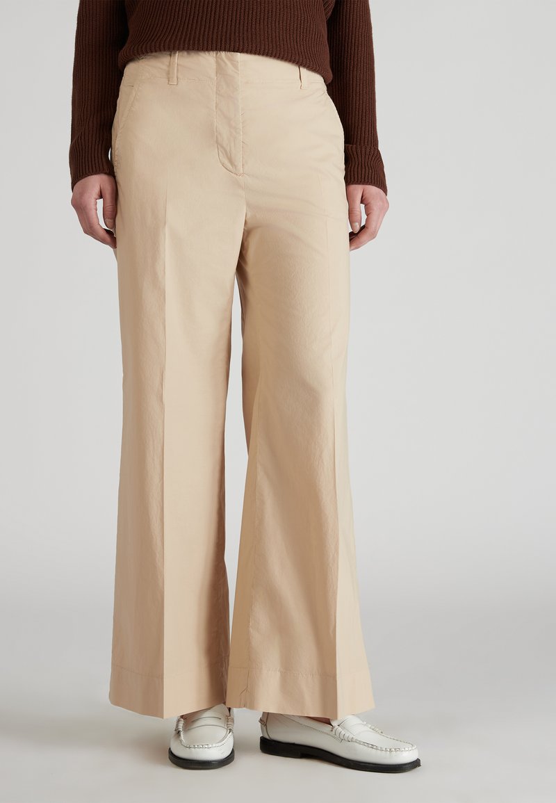 Persona con pantalones de pierna ancha beige, mocasines blancos y un suéter de canalé marrón, de pie contra un fondo claro y liso.