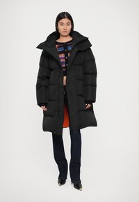 Abrigo tipo puffer negro con corte holgado, cuello sobredimensionado y cierres a presión. Presenta un forro interior naranja, combinado con pantalones de mezclilla oscuros.
