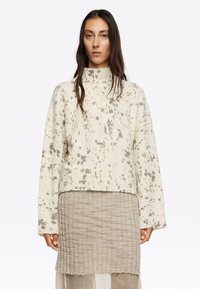 Maglione chiaro a collo alto, con vestibilità ampia e macchie marroni sparse; abbinato a una gonna lavorata a maglia beige.