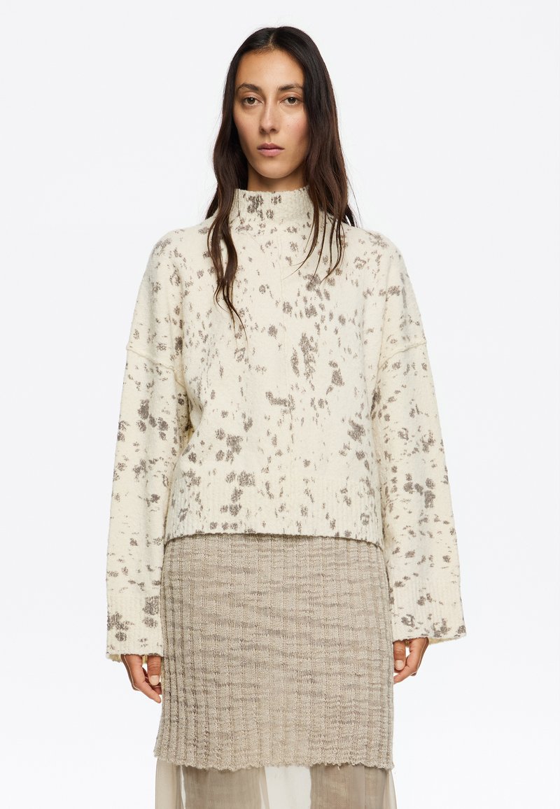 Maglione chiaro a collo alto, con vestibilità ampia e macchie marroni sparse; abbinato a una gonna lavorata a maglia beige.
