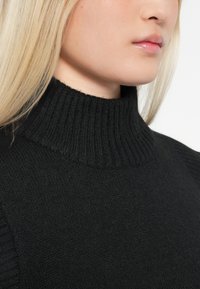 Zbliżenie na osobę o jasnych blond włosach, ubraną w czarny sweter z golfem, pokazujące szyję i dolną część twarzy.