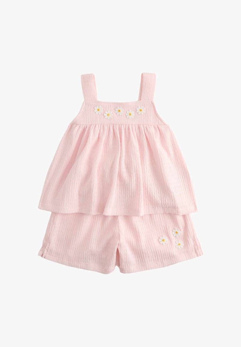 Mono corto sin mangas de color rosa claro con textura, con falda superpuesta fruncida y pequeños apliques de margaritas blancas en el pecho y los pantalones cortos.