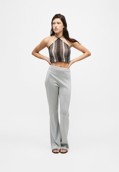 Jeune femme debout, les mains sur les hanches, portant un top court à bretelles avec des rayures verticales et un pantalon évasé gris taille haute.