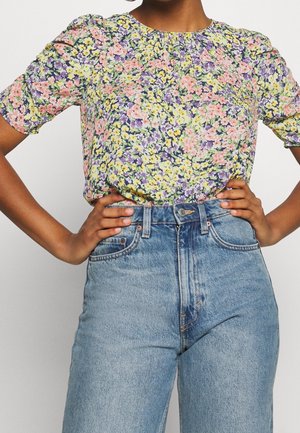 Person med hænderne på hoften iført en farverig blomstret kortærmet bluse stoppet ned i højttaljede lyseblå jeans.