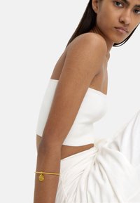 Biała, bezramiączkowa bluzka, materiał w kolorze cielistym, o gładkiej fakturze. Na nadgarstku znajduje się prosty złoty bransoletka z okrągłym zawieszką.