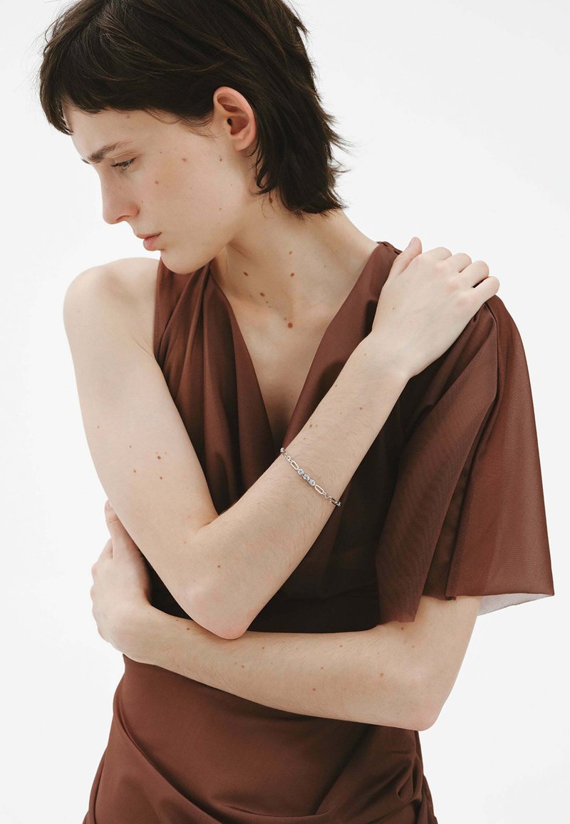 Vestido marrón sin mangas con un tejido suave y fluido. La modelo tiene el cabello corto y lleva una pulsera de plata en un brazo. Fondo claro.