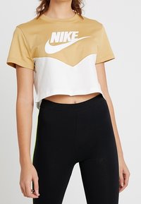 Avkortad Nike-t-shirt med ett design i guld och vitt färgblock, stor logotyp, rund halsringning och korta ärmar, paras ihop med svarta leggings.