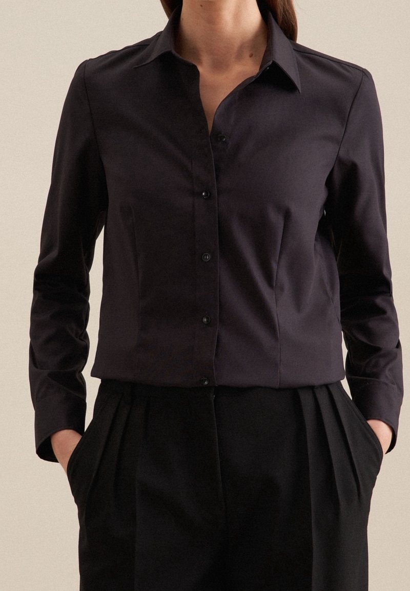 Chemise à boutons ajustée, de couleur sombre, avec col, manches longues et tissu lisse ; associée à un pantalon noir taille haute.