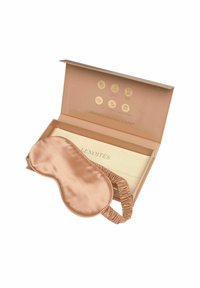 Saténová oční maska v barvě růžového zlata s elastickým páskem, umístěná v jasně béžové krabičce s zlatými ikonami a brandingem.