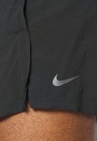 Pantalones cortos deportivos negros de tejido ligero con un brillo sutil. Presentan un logo plateado de Nike en la pierna izquierda y aberturas para bolsillos laterales.