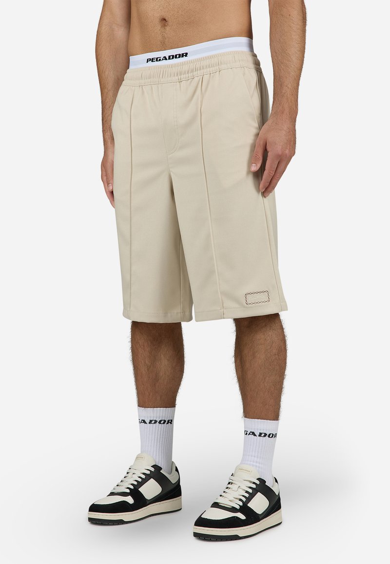 Shorts sportivi beige con vita elasticizzata e pieghe laterali, caratterizzati da un piccolo patch con logo vicino all'orlo. Tessuto misto cotone.