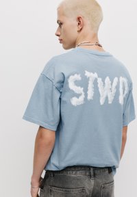 Tricou din bumbac de culoare albastru deschis, cu litere albe asemănătoare norilor "STWD" pe spate, croială lejeră, cu mâneci scurte și guler clasic.