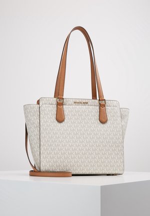 Witte en beige Michael Kors handtas met monogrampatroon, twee lange beige hengsels en een afneembare schouderriem op een wit voetstuk.
