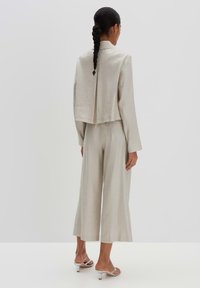 Lichtbeige twee-delige outfit met een korte blazer met een rugplooien en rechte broek. G gedragen met zilveren hoge hakken.