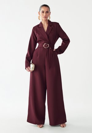 Femme portant une combinaison bordeaux ceinturée avec des jambes larges, tenant une petite pochette dorée et portant de grandes boucles d’oreilles dorées.