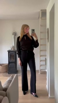 Blusa negra de manga larga con escote tipo ojo de cerradura, combinada con pantalones negros ajustados. Tejido suave con un corte ligeramente holgado. Zapatos negros de punta.