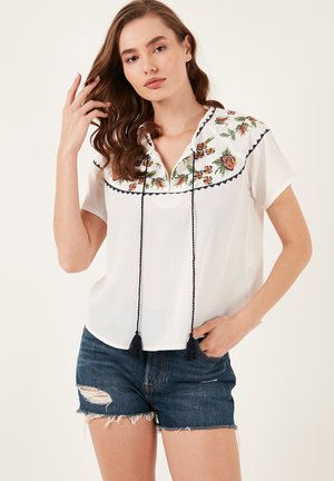 White short-sleeve blouse with floral embroidery around the collar, featuring a placket and black tassel accents. Paired with denim shorts.