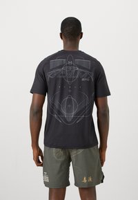 T-shirt noir avec un grand imprimé graphique géométrique blanc au dos, manches courtes, col rond et tissu léger.