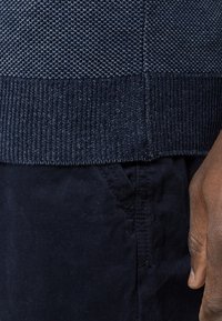Ourlet de pull texturé bleu foncé au-dessus de la poche d’un pantalon bleu foncé et du côté d’une main d’une personne à la peau brune.
