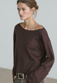 Maglia a maniche lunghe color marrone rossiccio con scollo ampio, vestibilità rilassata e trama a costine, abbinata a jeans scuri e cintura con fibbia d'argento.