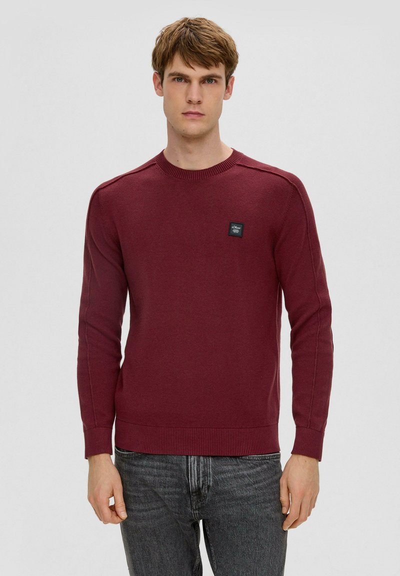 Burgunder gestrickter Pullover mit Rundhalsausschnitt, gerippten Ärmelbündchen und Saum sowie einem schwarzen Etikett auf der Brust, kombiniert mit dunkelgrauen Jeans.