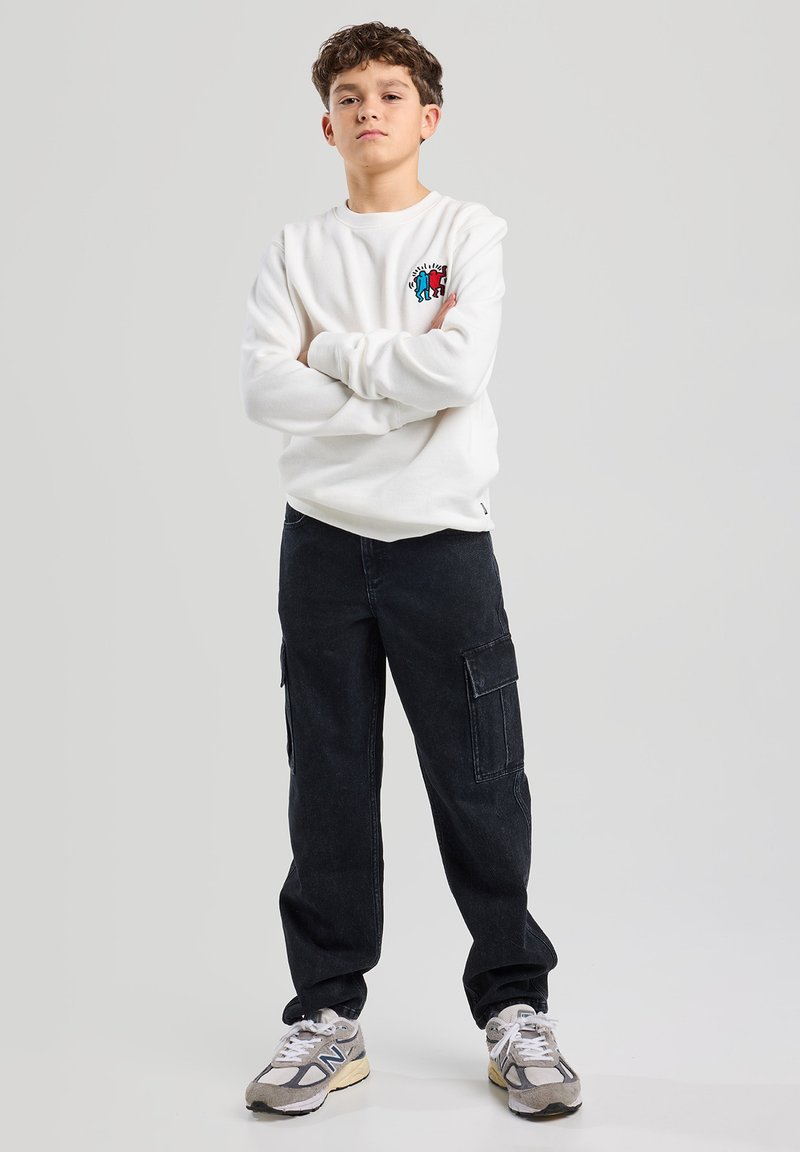 Felpa bianca con un dettaglio grafico colorato, abbinata a pantaloni cargo neri con tasche laterali e sneakers grigie. Sfondo semplice.