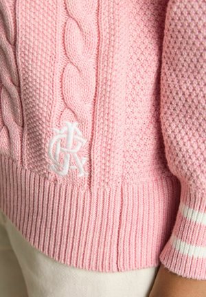 Rozā adīts džempera ar divām baltām svītrām pie ribveida apakšmalas un aprocēm, valkāts ar bēšām biksēm.