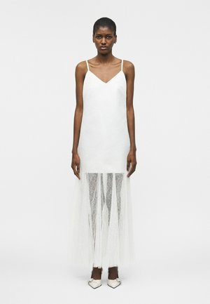 Mujer con un vestido blanco sin mangas con una superposición de encaje transparente que se extiende desde la mitad del muslo hasta el suelo, combinado con zapatos de tacón blancos de punta puntiaguda.