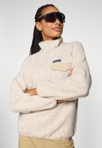 Fleece-Pullover in Hellbeige mit subtilen geometrischen Mustern, Stehkragen, Fronttasche und Druckknöpfen. Getragen mit übergroßen schwarzen Sonnenbrillen.