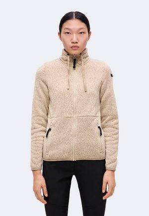 Kvinde iført beige fleecejakke med lynlås, høj krave og snore, kombineret med sorte bukser, stående foran en ensfarvet baggrund.