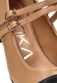 Zapato de cuero sintético beige con puntera negra, con correas cruzadas y hebillas metálicas. Logo dorado impreso en la plantilla.