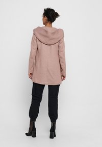 Cappotto di lana rosa chiaro con un grande cappuccio, caratterizzato da una linea dritta e maniche lunghe, abbinato a pantaloni cargo neri e stivaletti alla caviglia.