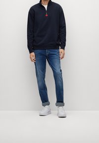 Felpa zip navy con un accento rosso, abbinata a jeans azzurri chiari e sneakers bianche, il tutto su uno sfondo neutro.
