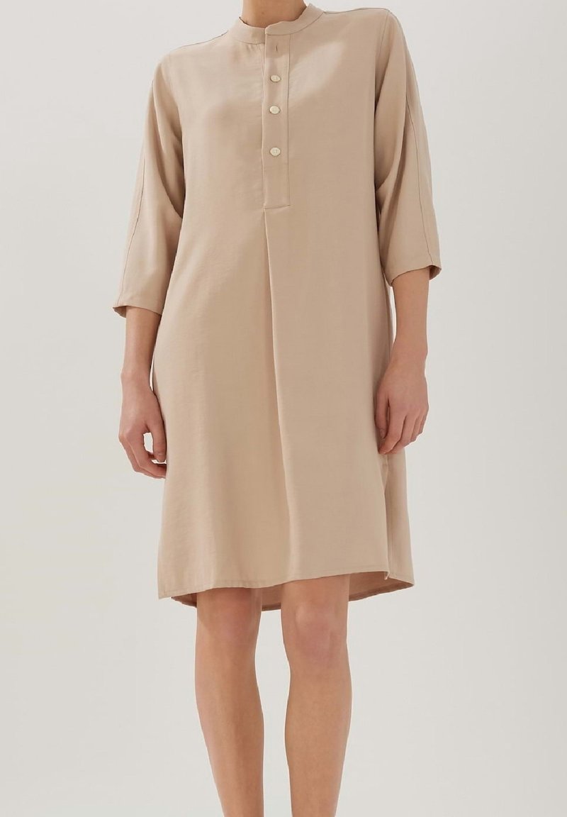 Personne portant une robe beige ample jusqu'aux genoux avec des manches trois-quarts et une patte de boutonnage sur un fond clair.