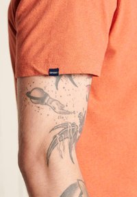Kortærmet orange T-shirt med blød tekstur og et lille blåt "Superdry" mærke nær manchetterne, tæt på en tattoveret arm.