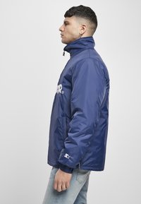 Tamsiai mėlynas vėjo skutimo švarkas su aukštu apykakle, užtrauktuku, elastinėmis rankovėmis ir šoninėmis kišenėmis. Priekį puošia baltas logotipas.