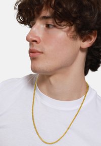 Collier en chaîne dorée avec des maillons carrés, suspendu à un t-shirt blanc. Fini brillant, épaisseur moyenne, il repose confortablement autour du cou.
