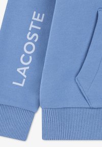 Φωτεινή γαλάζια φούτερ με ριμπ μανσέτες και τελείωμα, με έντονο λευκό λογότυπο "LACOSTE" τυπωμένο κάθετα στο μανίκι. Ομαλή υφή.