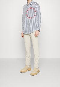 Pruhovaná košile na knoflíky s červeným grafickým potiskem, béžové kalhoty s rovnými nohavicemi a béžové boty. Košile má dlouhé rukávy a zaoblený lem.