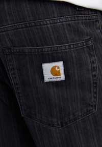 Schwarze Jeans aus Denim mit einer auffälligen Gesäßtasche, die ein quadratisches weißes Etikett mit einem gelben Carhartt-Logo zeigt. Subtile vertikale Textur.