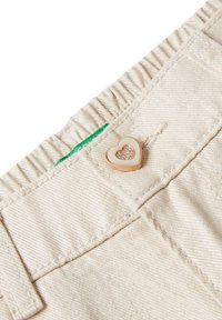 Zbliżenie na talię beżowych spodni z guzikem w kształcie serca ozdobionym cyrkoniami oraz małą zieloną metką.