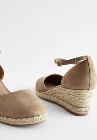Paire d'espadrilles compensées en daim beige avec brides autour de la cheville et semelles en jute tressé sur fond blanc.