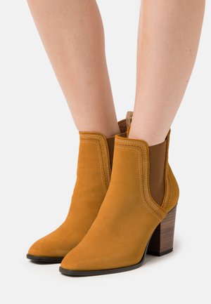 Tan suede ankle boots with a pointed toe and stacked wooden heel. Features elastic side panels and contrasting white stitching details.