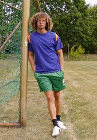 Athletic figure wearing a blue shirt with orange accents and green shorts. Stands near a goalpost, grassy field in background.