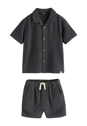 Jeune garçon portant une chemise gris foncé à manches courtes boutonnée et un short assorti, debout devant un fond beige neutre.