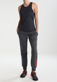 Zwarte mouwloze sporttanktop met reliëflogo, gecombineerd met grijze taps toelopende joggers met rode branding, en zwarte sportschoenen.