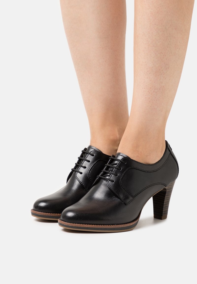 Chaussures à talons en cuir noir avec bout arrondi, laçage à l'avant et panneaux latéraux texturés. Le talon est de forme carrée avec des accents en bois.
