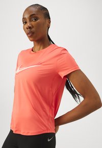 Camiseta atlética de Nike en un vibrante color coral, con cuello redondo, mangas cortas y un logo de swoosh blanco en el frente. Tejido suave y ligero.
