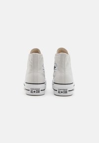 Baskets montantes en toile blanche avec des semelles en caoutchouc noir, présentant un logo circulaire sur le côté extérieur et deux bandes noires sur la semelle.