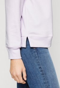 Felpa viola chiaro con polsini a coste, tessuto leggermente strutturato e orlo laterale spaccato, abbinata a jeans di denim blu.